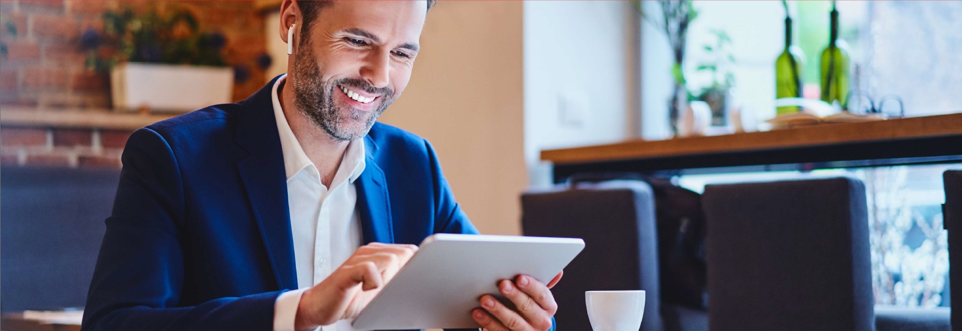 A man in a business suit with earpods in is smiling while using a tablet in a coffee shop